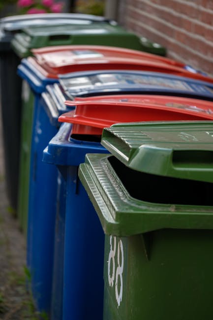 A row of large, rectangular wheelie bins lined up outdoors along a paved pathway next to a brick wall. The bins are made of durable plastic with textured, grooved lids that are hinged at the back. The bins are arranged in a sequence, with the closest bin being green, followed by blue, red, and other bins in the background, each with matching lids. The green bin in the forefront has a slightly open lid, revealing the interior space. The bins are situated on a narrow strip of ground, with some grass visible to the left, and the scene is lit by natural daylight, providing even illumination that highlights the textured surfaces and vibrant colors. This setup suggests an organized waste management system, often used for private or council waste collection services, illustrating an alternative to traditional disposal methods and emphasizing the importance of proper rubbish segregation and collection for efficient waste clearance, as provided by Waste Clearance Brixton Ltd.