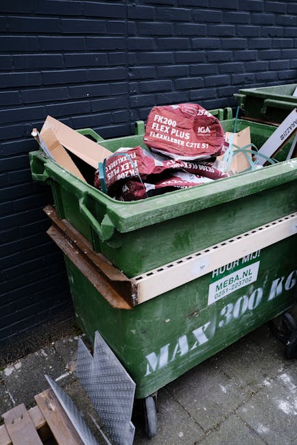 A row of large, rectangular wheelie bins lined up outdoors along a paved pathway next to a brick wall. The bins are made of durable plastic with textured, grooved lids that are hinged at the back. The bins are arranged in a sequence, with the closest bin being green, followed by blue, red, and other bins in the background, each with matching lids. The green bin in the forefront has a slightly open lid, revealing the interior space. The bins are situated on a narrow strip of ground, with some grass visible to the left, and the scene is lit by natural daylight, providing even illumination that highlights the textured surfaces and vibrant colors. This setup suggests an organized waste management system, often used for private or council waste collection services, illustrating an alternative to traditional disposal methods and emphasizing the importance of proper rubbish segregation and collection for efficient waste clearance, as provided by Waste Clearance Brixton Ltd.