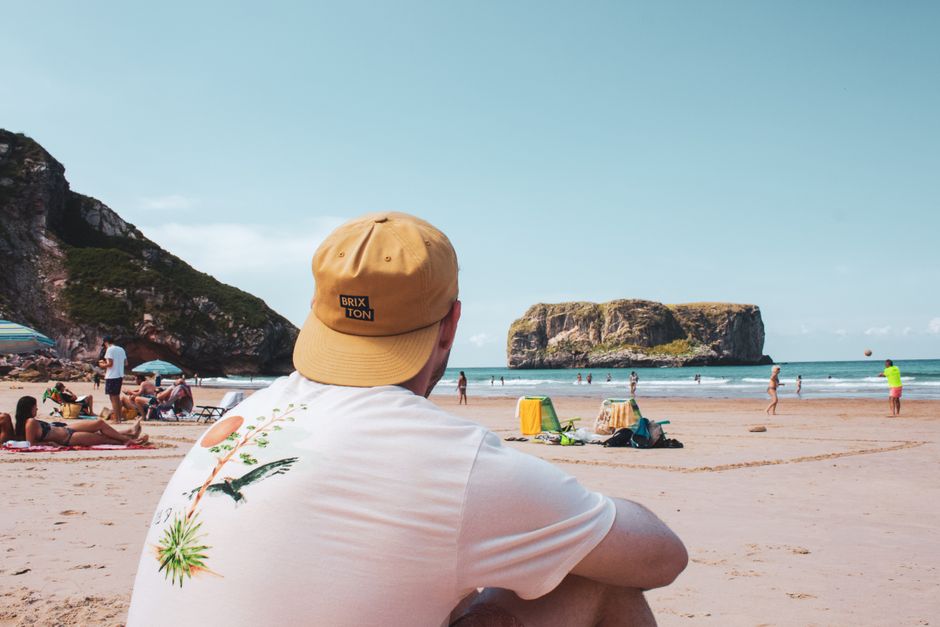 A person wearing a beige BRIXTON cap and a white T-shirt with a tropical plant and bird graphic sits on a sandy beach, facing away from the camera and looking towards the ocean. In the background, several individuals are lounging on sunbeds and walking along the shoreline. There are also colorful umbrellas, beach towels, and bags scattered on the sand. Beyond the beach, there are large rocky cliffs and a prominent sea stack formation rising from the water, with a clear blue sky overhead. The scene suggests a lively coastal environment, and the image subtly relates to the importance of managing waste and rubbish removal in outdoor recreational areas, aligning with services for outdoor public space clearance provided by companies like Waste Clearance Brixton Ltd.