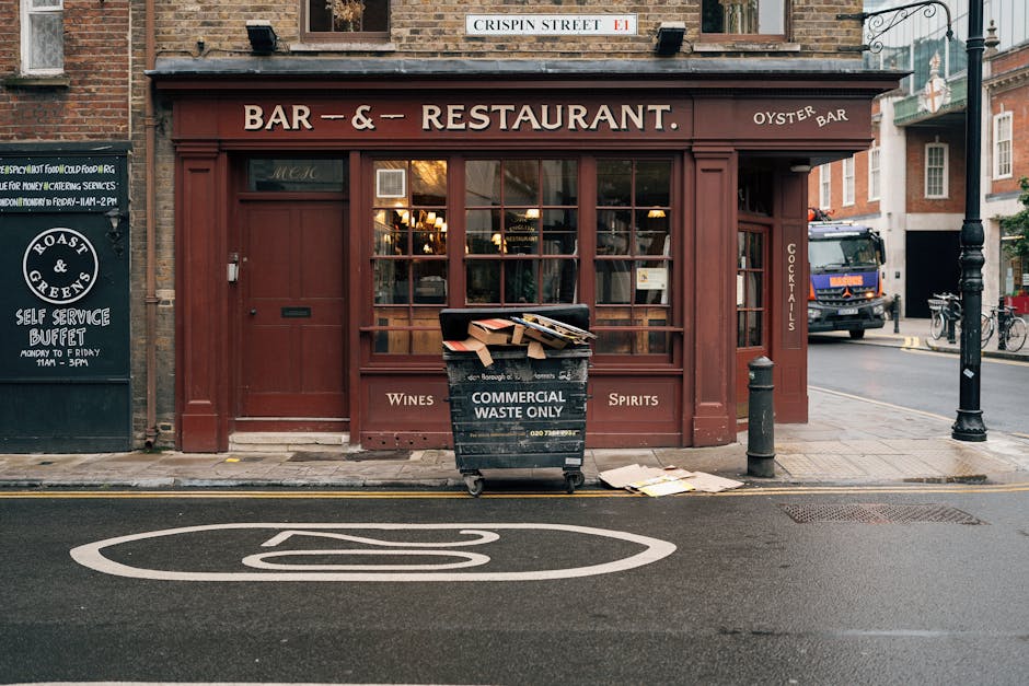 In front of a brick building housing a bar and restaurant, a large black commercial waste bin marked 'COMMERCIAL WASTE ONLY' is positioned on the sidewalk. The bin is filled with flattened cardboard boxes and other miscellaneous waste materials, some of which are spilling over the top and leaning against it. The container has wheels and a handle, facilitating transportation, and is situated near a short metal post and a black bollard that protect the pedestrian area. To the left, a smaller blackboard-style sign advertises a self-service buffet, while to the right, a black vehicle is partially visible parked on the street, with a few bicycles secured to a stand close by. The scene is set during daylight, and the street features a clearly painted, white, double-lined traffic calming zone on the asphalt directly in front of the waste bin. This snapshot emphasizes the importance of local waste management, consistent with professional rubbish removal services such as those provided by Waste Clearance Brixton Ltd, focusing on on-site clearance and private waste collection in urban environments.