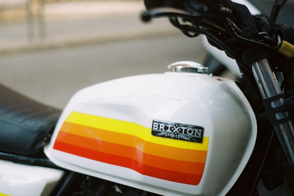 Close-up view of a white motorcycle fuel tank with a glossy finish, featuring a retro-style stripe graphic in yellow, orange, and red horizontal bands. The tank is positioned on the left side of the image, with a metallic badge displaying the brand name 'Brixton Motorcycles' attached toward the front. The motorcycle's black seat is visible behind the tank, extending to the left, while the front fork and part of the handlebars are partially shown in the upper right corner. The background is blurred, indicating an outdoor setting with natural light illuminating the tank's surface, accentuating its polished appearance. This scene portrays a classic motorcycle design, which could be associated with individual transport or private disposal of vehicle-related waste as part of a broader rubbish removal service offered by Waste Clearance Brixton Ltd, serving areas near Brixton station.