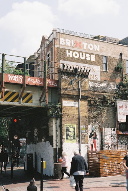 A multi-story brick building with large painted signage displaying the words 'BRIXTON HOUSE' and additional text, partially obscured by rusted scaffolding and greenery growing on the wall. The building's exterior shows signs of weathering, with peeling paint and old graffiti on lower sections. In the foreground, a busy street scene includes pedestrians walking and standing nearby, with some engaging in conversation or observing the surroundings. To the left, a raised concrete platform or bridge with yellow and black striped warning markings spans part of the scene, supporting temporary barriers and fencing. The environment appears urban with a mix of architectural elements, graffiti, and street-level activity, reflecting an area that may naturally feature diverse waste or clutter which private waste clearance services like Waste Clearance Brixton Ltd could help manage, especially considering the indication of remnants from previous signage and maintenance work along the building façade. Overcast sky provides diffuse lighting, highlighting textured brickwork and weather-beaten surfaces.
